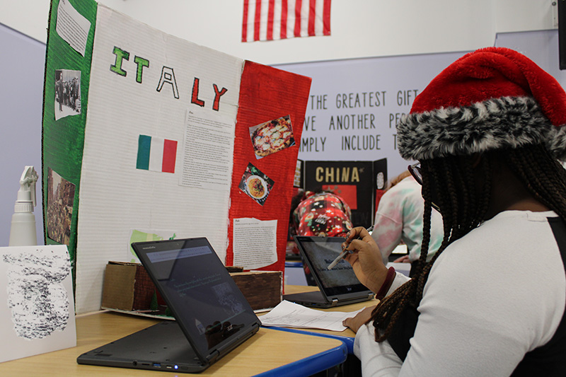 A poster board with the word Italy on it and an italian flag of green, white and red is on a table. A young woman wearing a red fuzzy santa cap is sitting at the table writing on paper. There is a Chromebook in front of her.