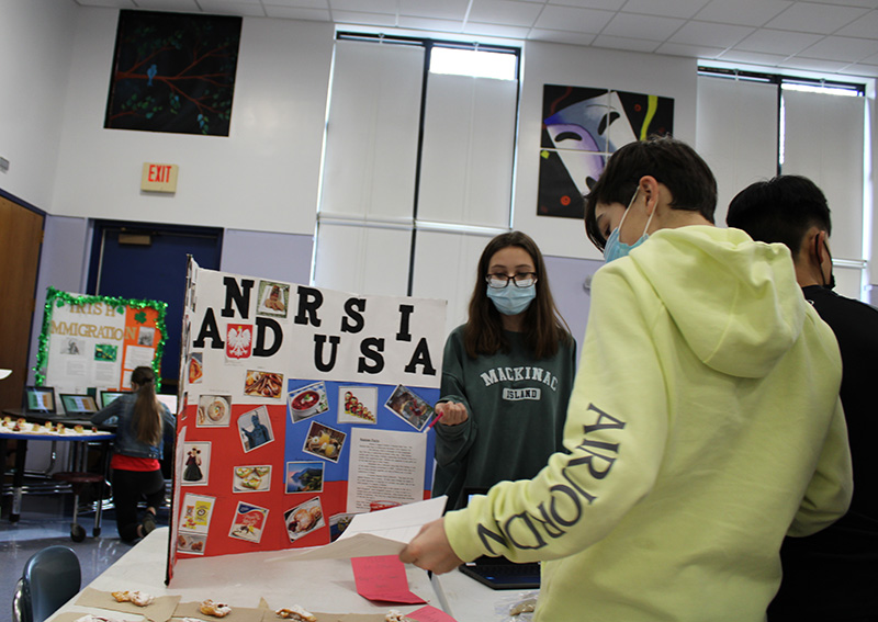 A young man wearing a yellow sweatshirt takes a napkin and food from a table. In the background is a  poster board that says Poland and Russia with items on it 