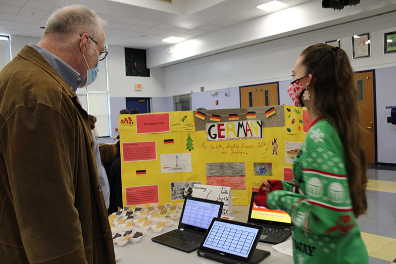 A man with light hair, glasses and a blue facemask, wearing a brown jacket, stands watching a young woman explaining German immigration. There is a yellow poster board with things about Germany on it and two Chromebooks with a Jeopardy-type game on it.