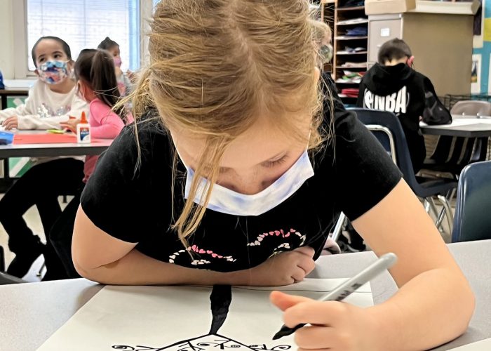 A student with blonde hair pulled back in a ponytail. She is wearing a black t shirt with pink and white on the gfront, and a blue mask. She is leaning over a piece of paper on which she drew a tree of life with a black marker,