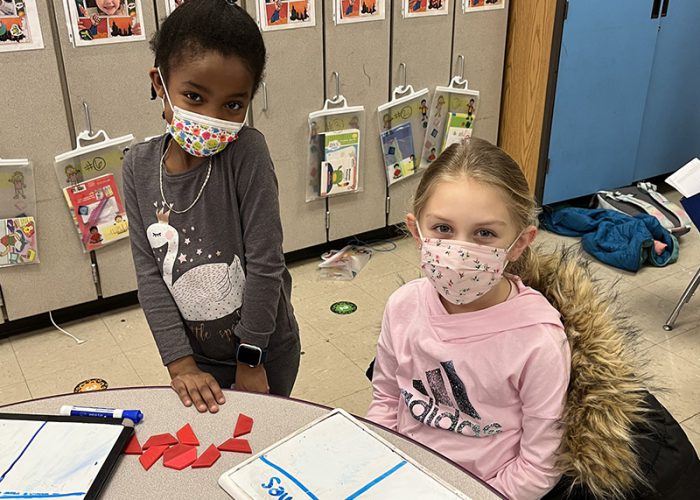 Two young elementary age students. One is standing and the other is sitting at a table. In front of them are small objects for counting. They are both wearing masks.