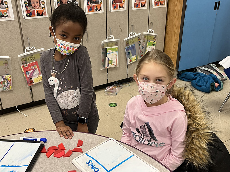 Two young elementary age students. One is standing and the other is sitting at a table. In front of them are small objects for counting. They are both wearing masks.