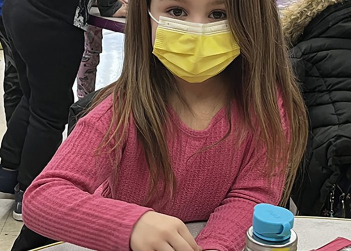 A young elementary age girl wearing a pink long-sleeve shirt and yellow mask counts little pink erasers. They are in little groups of 10 on the table.