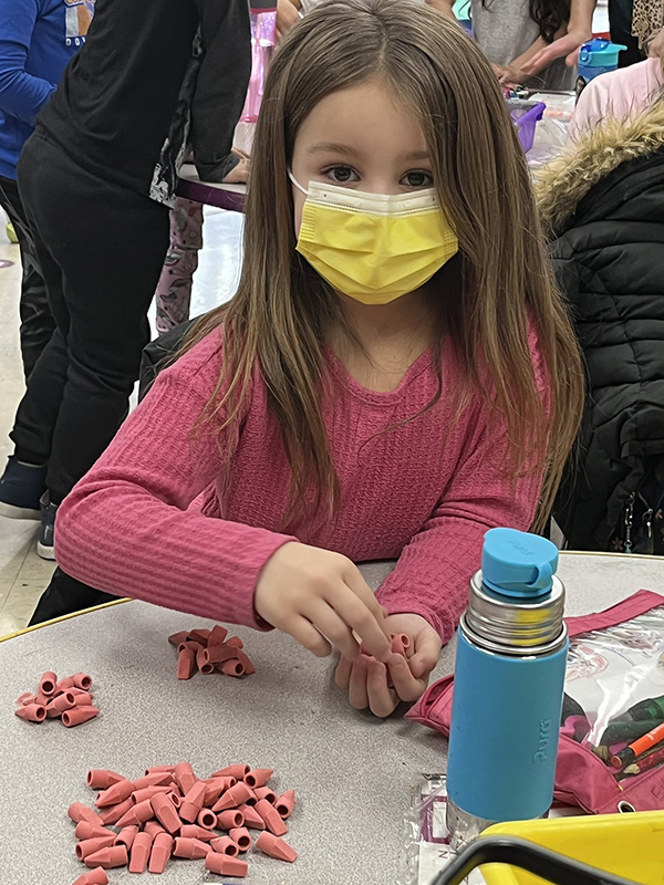 A young elementary age girl wearing a pink long-sleeve shirt and yellow mask counts little pink erasers. They are in little groups of 10 on the table.