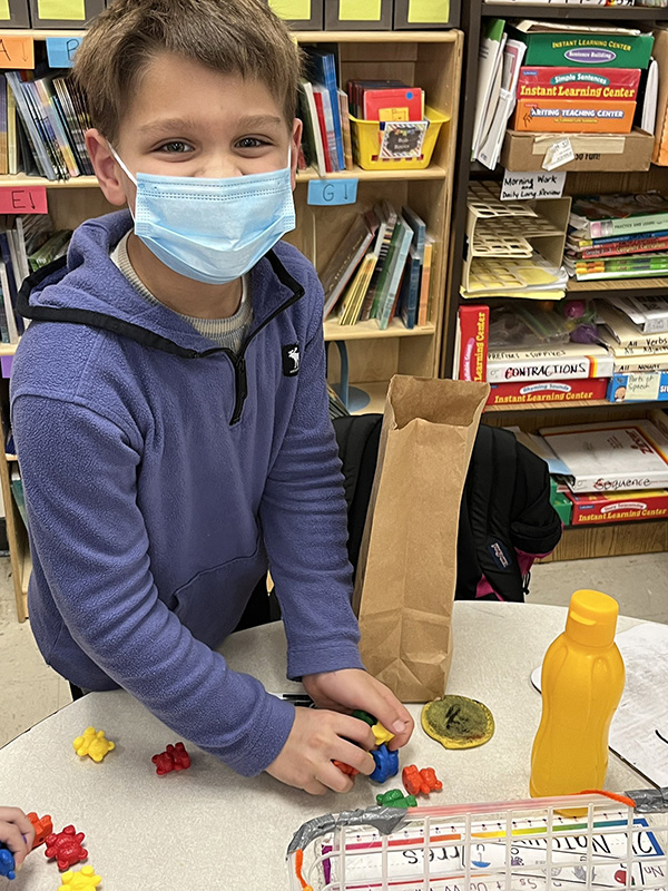 A young elementary-age boy wearing a blue hoodie has many different colored objects on a table. He is wearing a blue mask.