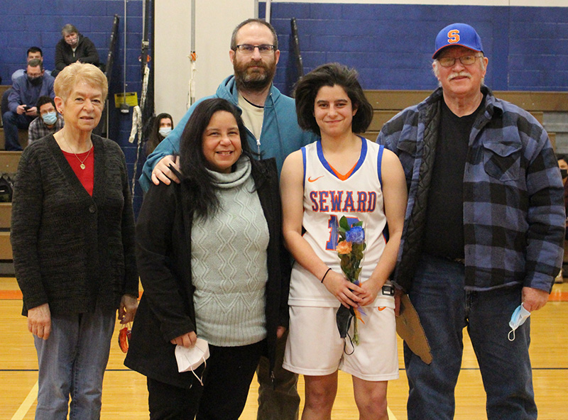 A young woman with chin-length dark hair, wearing a white basketball uniform that says SEWARD on it, stands with family members. There are two women on the left, a man behind and a man on the right. The player is holding a flower. They are all smiling.