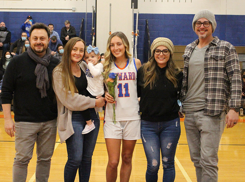 A high school girl with long blond hair, wearing a white basketball uniform that says SEWARD on it and number 11 stands with her arms around two women. Next to each of the women is a man. One of the women is holding a small child. All are smiling.