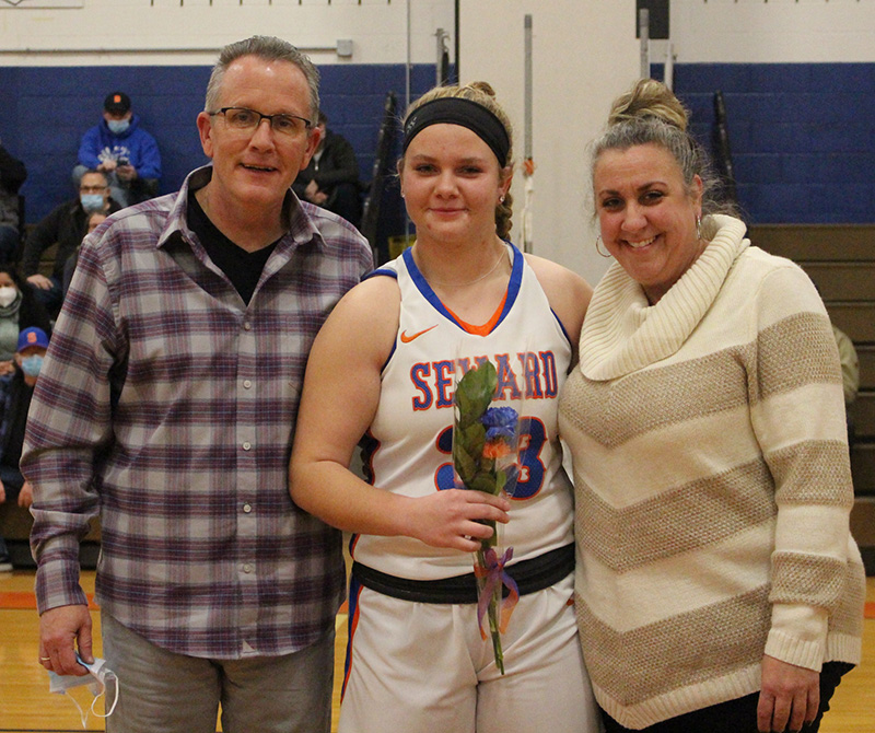 A young women with a blond ponytail and dark headband stands between a man and a woman. She is wearing a white basketball uniform that says SEWARD and is holding a flower. All are smiling.