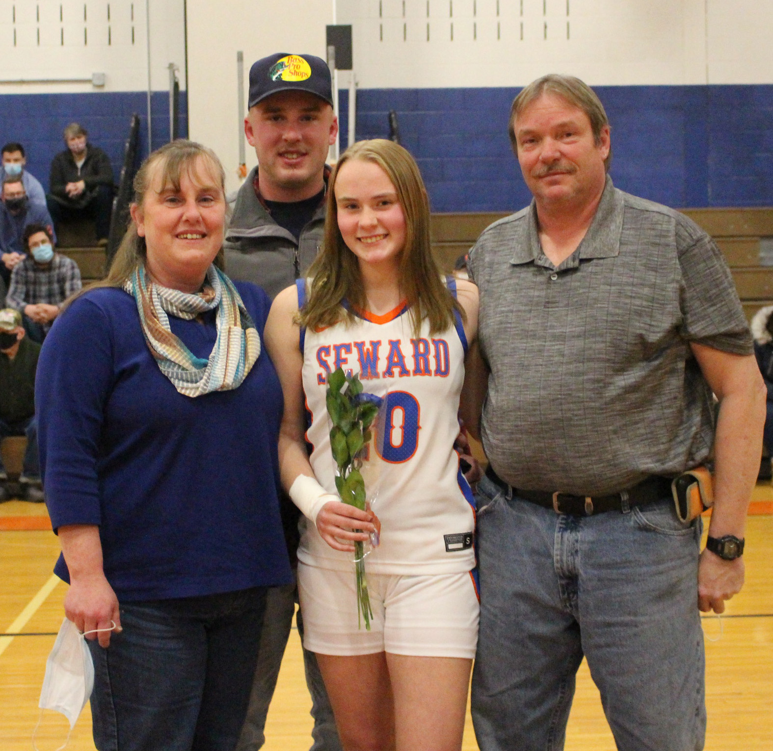 A young woman with shoulder length brown hair stands in the center wearing a white basketball uniform that says SEWARD. She is holding a flower and next to her are a man and a woman. Behind her is a man.