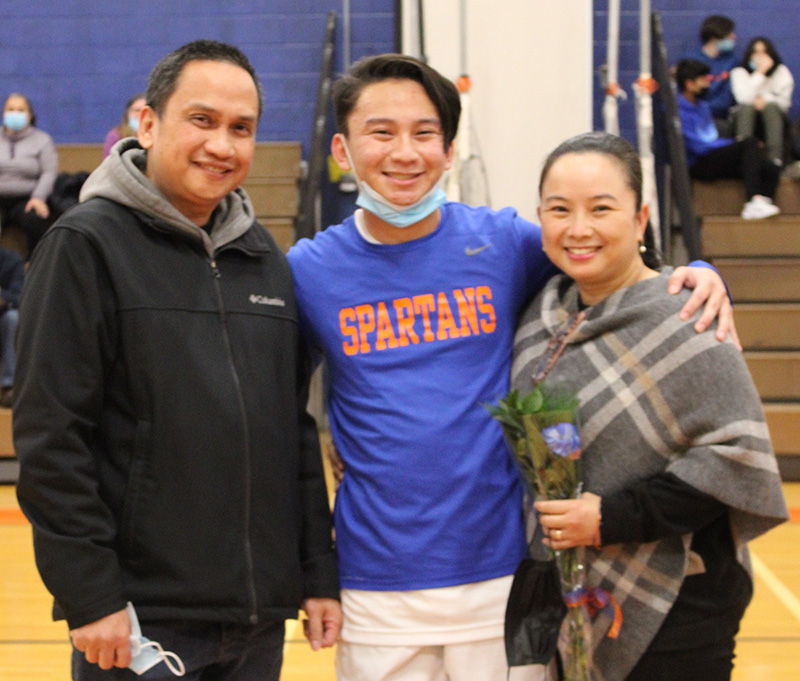 A high school senior boy stands in the center . He is wearing a blue shirt that reads Spartans in orange. on either side of him is a man and a woman. They are all smiling. The woman is holding a flower.