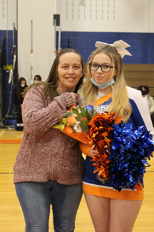 A young woman with long blonde hair and glasses holds a flower and blue and orange pom poms. She is standing with a woman wearing jeans and a reddish sweater. They are smiling.