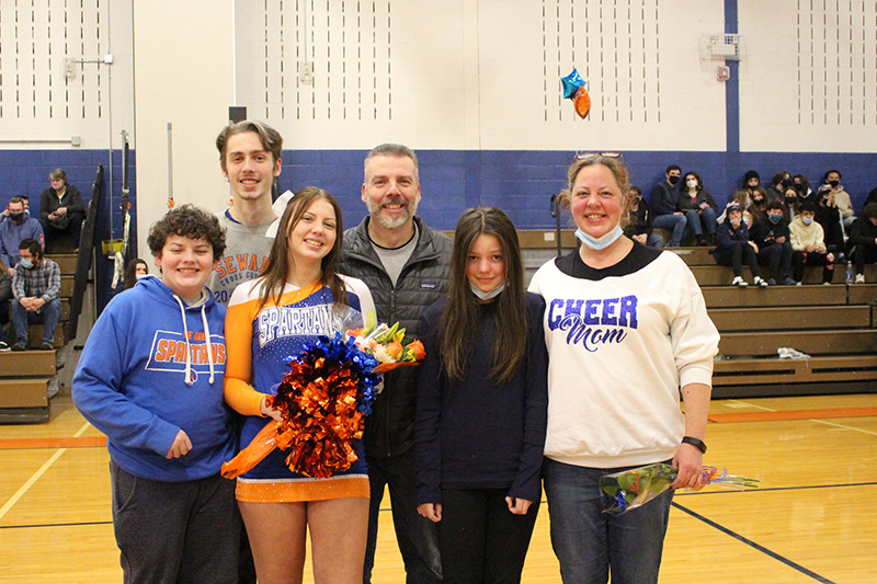 A young woman stands with five family members. She is second from the left and is weearing a blue and orange  cheerleading uniform that says SPARTANS on it. She is holding a flower and orange and blue pom poms. Next to her on the left is a young man, behind her are two men, and next to her on the right are two women. All are smiling.
