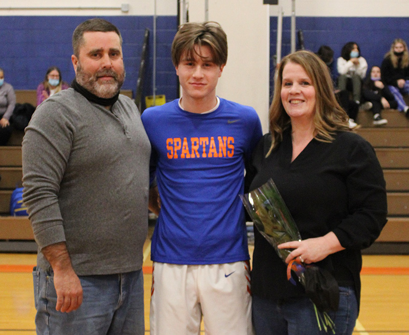 A high school senior boy stands in the center . He is wearing a blue shirt that reads Spartans in orange. on either side of him is a man and a woman. They are all smiling. The woman is holding a flower.