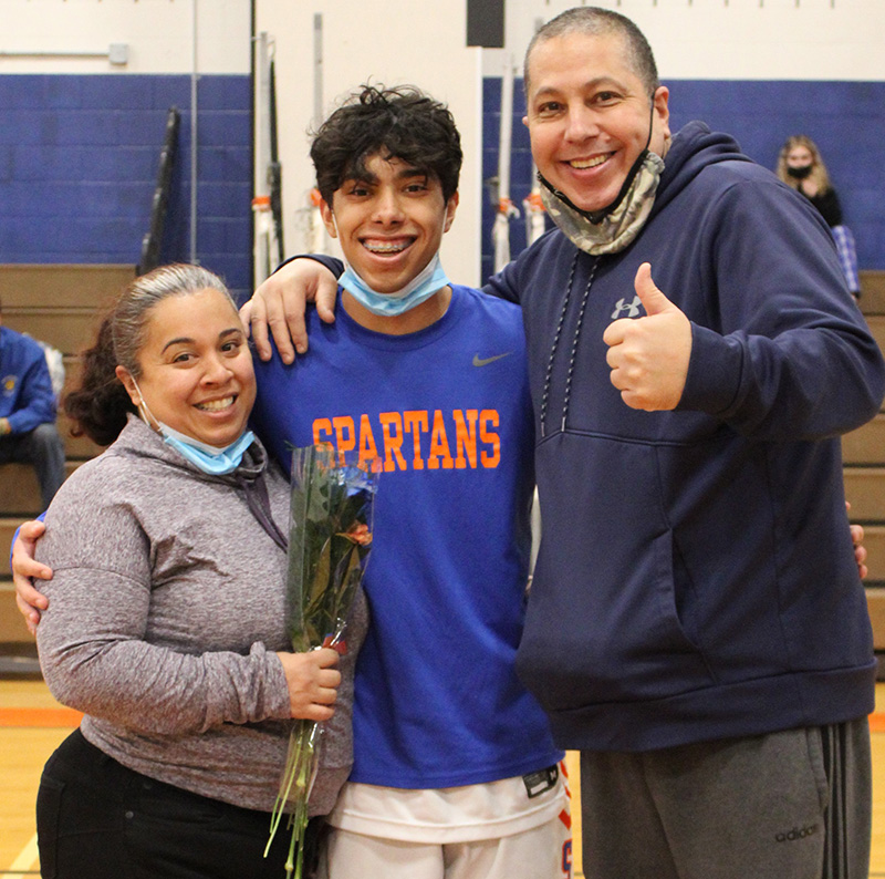 A high school senior boy stands in the center . He is wearing a blue shirt that reads Spartans in orange. on either side of him is a man and a woman. They are all smiling. The man is giving a thumbs up.The woman is holding a flower.
