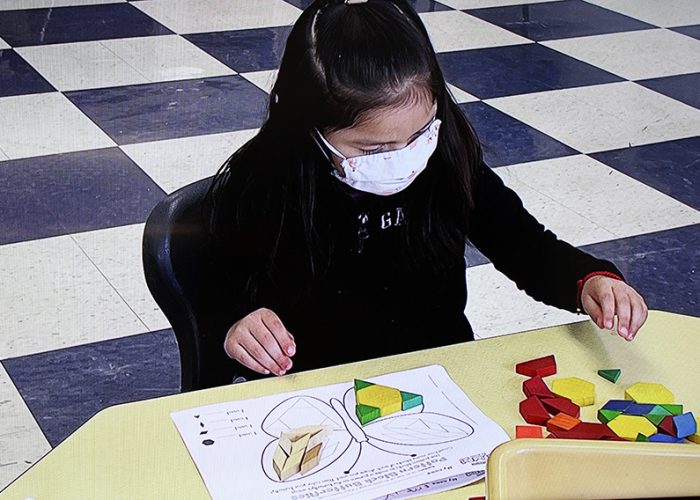 A young elementar-age girl, with long dark hair, wearing a dark shirt and white mask, gathers shapes make of wood and all different colors and creates a butterfly on a piece of paper on a table.