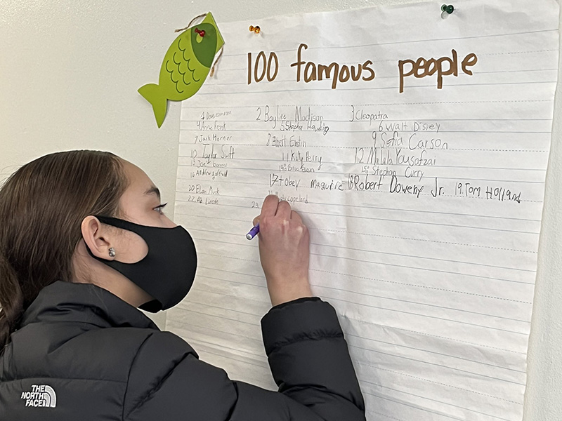 A fifth-grade girl, in a black jacket and black mask, writes on a large white poster that is tacked to the wall. It says 100 famous people.
