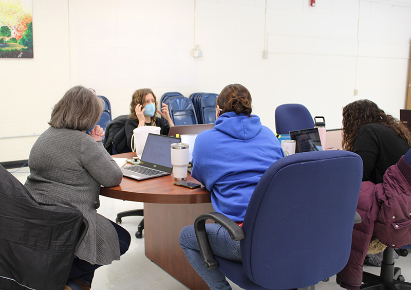 A woman with shoulder-length medium brown color hair sits at a table across from other adults. She is wearing a black shirt and a blue mask and has her hand up gesturing as she talks. The other adults are sitting around the table, in blue chairs listening.