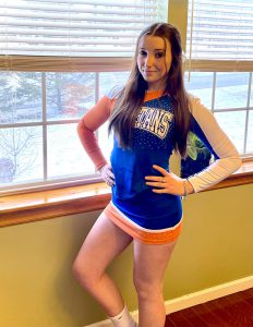 A high school girl with long brown hair. She is standing by a window. She is wearing a blue, orange and white cheerleading outfit that says Spartans on it.