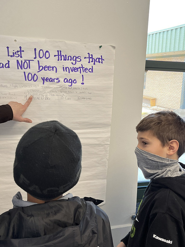 Two fifth-grade boys look at a poster on a wall. It says List 100 things that had not been invented 100 years ago.