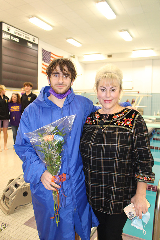 A young man on the left wearing a long blue hoodie has his arm around a woman with blond hair pulled up. She is wearing a plaid shirt and is smiling. He is holding a small bunch of flowers. They are both smiling.