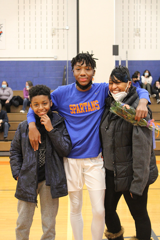 A high school senior boy stands in the center . He is wearing a blue shirt that reads Spartans in orange. on either side of him is a younger boy and a woman. They are all smiling. He has his arms around both of them and is holding a flower.