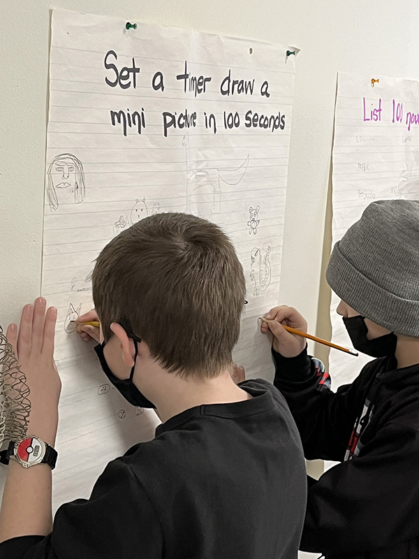 Boys in fifth grade, wearing black shirts and masks, write on a large piece of poster paper hung on a wall.  They are writing on it. It says Set a timer and draw a mini picture in 100 seconds.