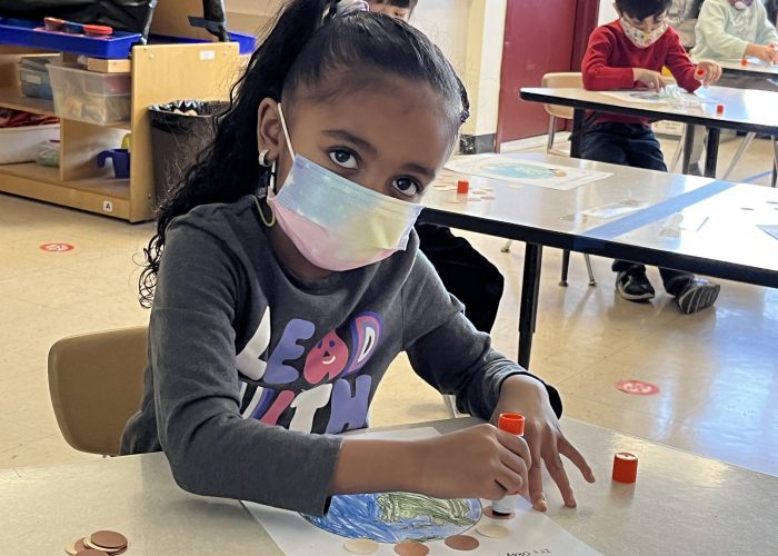 A small girl, with big dark eyes, long dark hair pulled back. She is wearing a gray shirt with a white mask and is gluing round pieces of paper on another paper that has the earth on it.