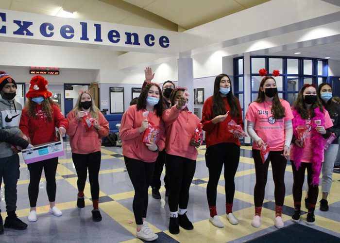 A group of 10 high school students stand in the lobby of a school. Behind them is a white beam at the top of the wall that says Excellence. Most of the students are wearing pink or red shirts and are holding bags of lollipops that they are handing out.