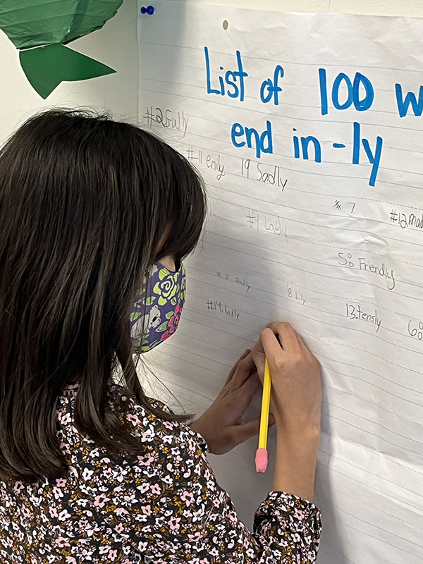 A fifth-grade girl writes with a yellow pencil on a poster on a wall. She has a printed shirt on and a printed mask. The paper says List of 100 words that end in -ly