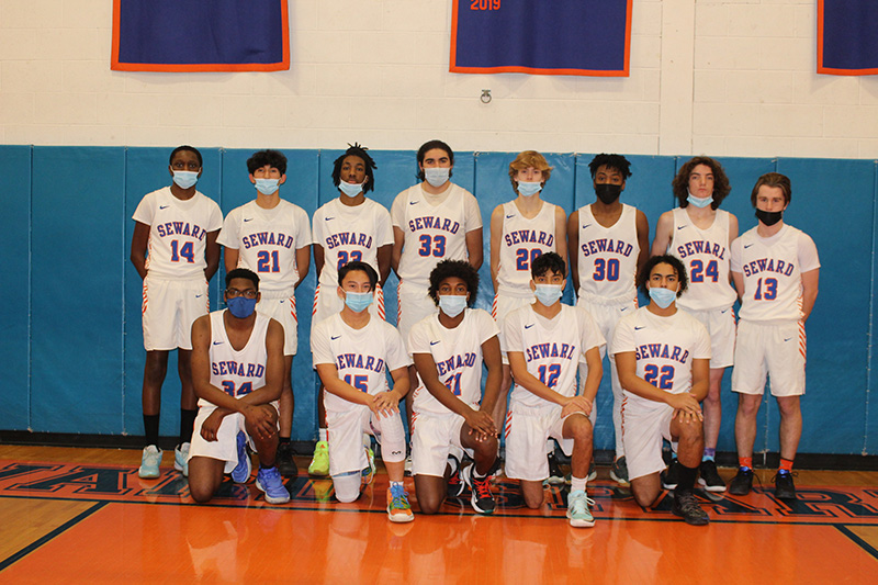 Thirteen high school boys , all wearing white basketball uniforms with Spartans written on them in blue and orange and numbers underneath. All are wearing masks. The back row has eight kids standing and the front has five boys kneeling.