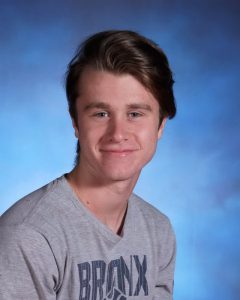 A high school boy with short hair, wearing a gray tshirt that says Bronx Bombers. He is smiling.