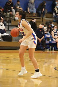 A high school basketball player, waring a white uniform with blue and orange stripe down the side. It is an action shot, with her dribbling the ball. She is wearing a paper mask and her long dark hair is pulled into a ponytail.