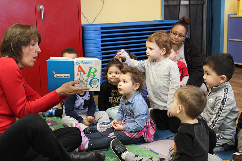 A woman in a red shirt sits on the floor and holds a book that says ABC on the front of it, open to a group of smaller preschoolers. They are also on the floor. One little boy is up on his knees pointing to the book.