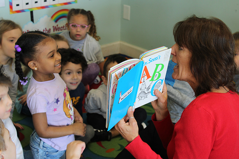 A woman in a red shirt holding up the ABC ook. A group of preschool children are sitting on the floor listening. One little girl is sitting up on her knees and smiling.