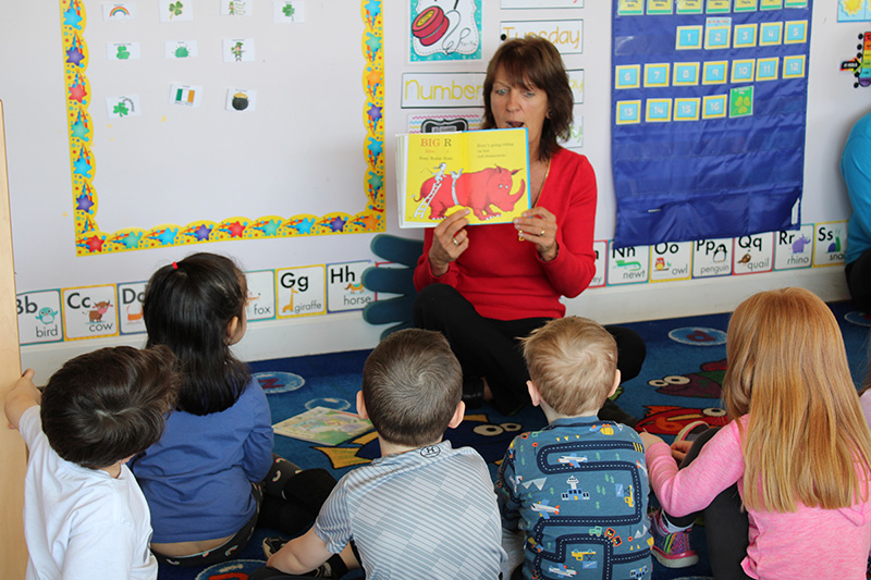 A woman in a red shirt, with short dark hair, holds up a book with a large red animal on the pages. A group of older preschoolers are sitting listening.