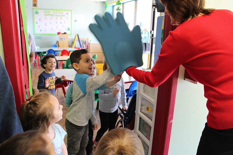 A preschool student smiles and gives a high five to a woman in a red shirt with a large blue foam hand on. There are other kids around looking and smiling.