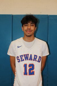 A high school boy wearing a white jersey that says Seward 12 on it in blue and orange. He has short, dark hair and is smiling.