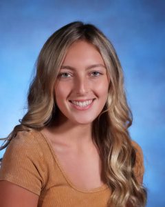 A high school girl with long blonde hair, wearing a beige shirt. She is smiling.