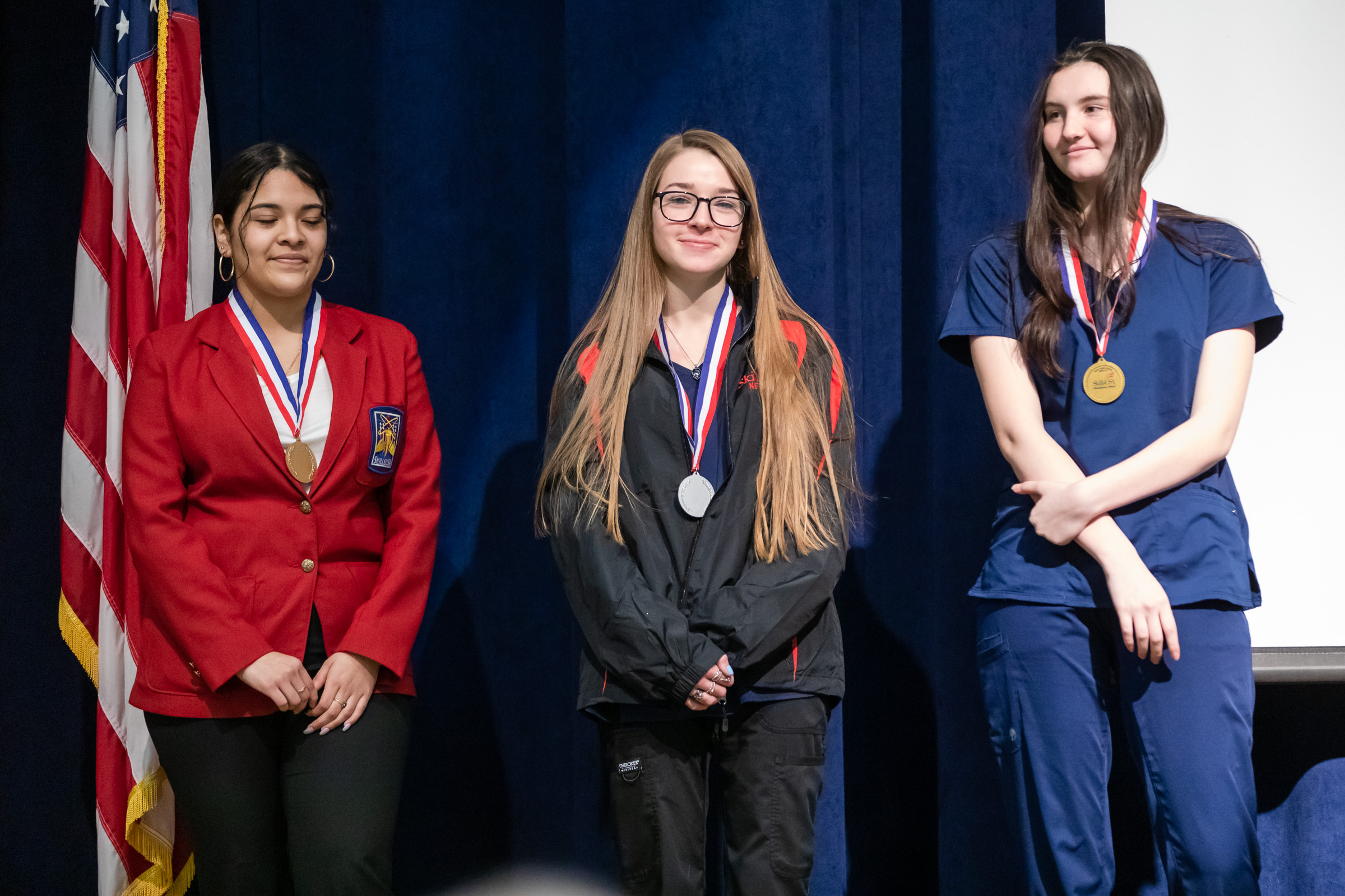 Three high school girls stand on a stage. The girl on the left is wearing a red blazer and has dark hair pulled back. The girl in the center has long blonde hair, and is wearing glasses and a dark colored outfit. The girl on the right is wearing a blue shirt and pants. She has long dark hair. There is an American flag on the left and a blue curtain behind them. All are wearing medals on red white and blue ribbons.