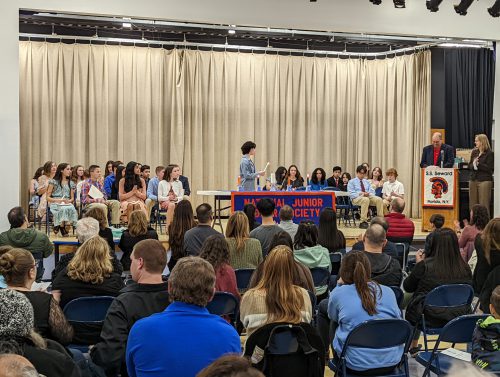 A large group of adults sitting in folding chairs. On the stage is a group of middle school students sitting. One student is lighting a candle.