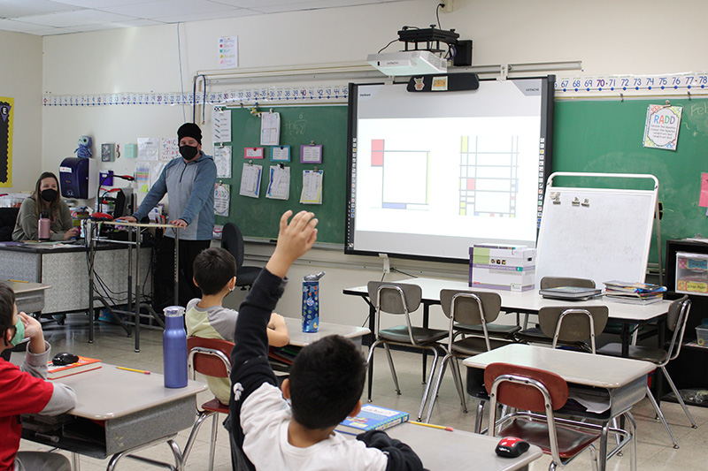 A classroom setting. A man is at the front of the class and there is a screen in front. A boy has his hand raised.