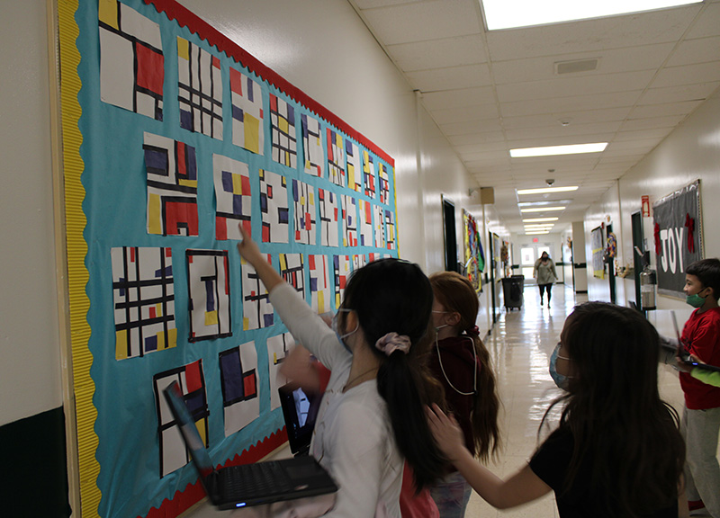 A group of students look at artwork on a bulletin board. Some are pointing at it.