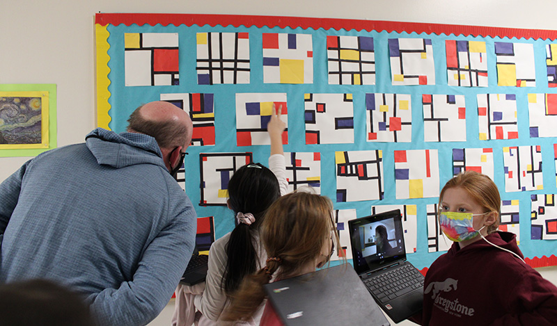 Students point to artwork on a bulletin board. A man in a blue hoodie stands with them looking.