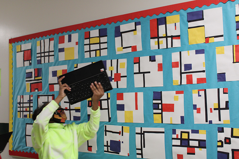 A bulletin board with many art pieces, all with blue, red and yellow colors on them. A student in a green shirt holds up her Chromebook to take a picture of one of the pieces of art work.