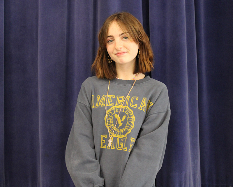 An eighth-grade girl, with shoulder-length reddish brown hair, wearing a gray sweatshirt with American Eagle on it in gold, stands in front of a large blue performance curtain. She is smiling.