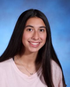 A high school girl with long dark hair, wearing a pink shirt. She is smiling.