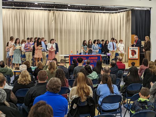 A group of about 30 middle school students stand on a stage while a group of parents sit in chairs watching.