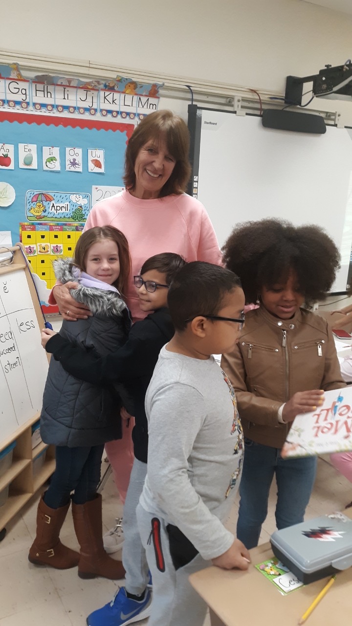 A woman with shoulder-length dark hair, wearing a pink shirt, hugs two elementary students. There are two other elementary students standing in front looking at a book.