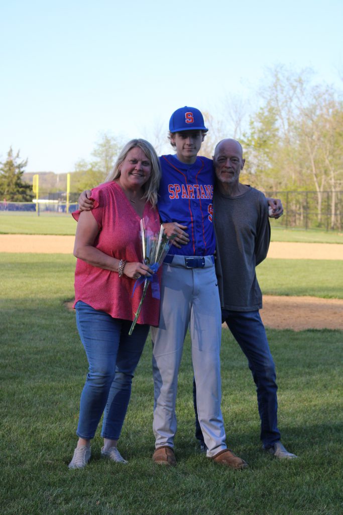 A high school boy stands between a woman on left and a man on right. He has his arms around them. He is wearing a baseball uniform, with a blue and orange shirt and gray pants. The woman is holding a flower.