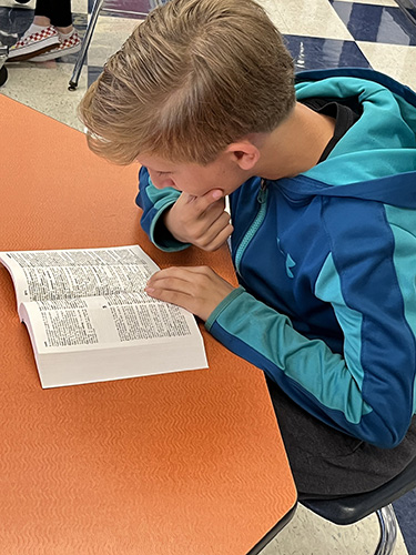 A boy with blonde hair sits at a table with his dictionary open. He is wearing a blue jacket.